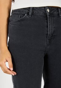 Schwarze Jeans aus Denim haben eine hohe Taille, eine schmale Passform und dezente Nähte. Der Stoff weist eine leichte Textur auf, und eine kleine Vordertasche ist sichtbar.