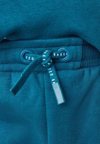 Los pantalones de chándal en color teal presentan una textura suave, un cordón ajustable con ojetes azules y una etiqueta de la marca impresa en el cordón.