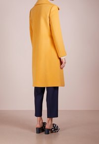 Cappotto di lana giallo con colletto strutturato, design fino al ginocchio e maniche lunghe, abbinato a pantaloni blu navy e mule a tacco largo con motivo.