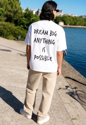 Man met zwart haar en zonnebril staat bij het water, draagt een wit overhemd met de tekst "DREAM BIG ANYTHING IS POSSIBLE" en beige broek.