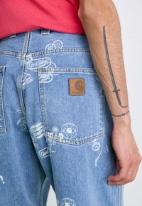 Jeans de mezclilla azul claro con patrones impresos en blanco, con un parche de cuero marrón con el logo y múltiples bolsillos en un diseño de corte relajado.