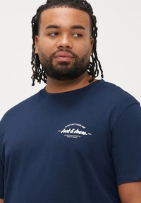 Μπλε σκούρο μπλουζάκι από βαμβάκι, με στρογγυλή λαιμόκοψη και κοντά μανίκια. Στο μπροστινό μέρος υπάρχει λευκό κείμενο και λογότυπο.