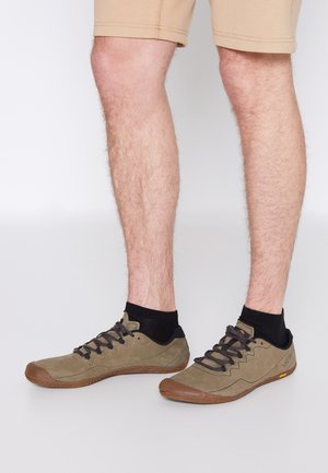 Gambe con pantaloncini beige, calzini neri alla caviglia e scarpe basse verde oliva con lacci e suole marroni su sfondo chiaro.