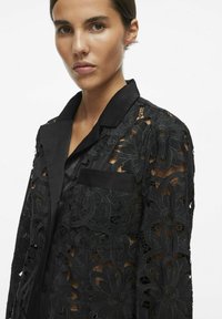 Chemisier en dentelle noire avec motif floral, texture transparente, col classique et poche poitrine unique. Il présente des détails riches et un design élégant.