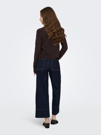 Dunkelbrauner figurbetonter Pullover kombiniert mit dunkelblauen Jeans mit weitem Bein. Die Jeans haben kontrastierende Nähte und einen gekürzten Saum. Geschlossene schwarze Schuhe.