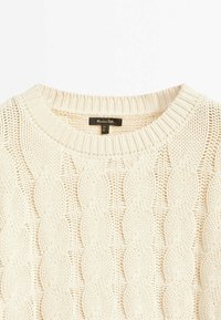 Pull en tricot de couleur crème avec un motif en coton et un col côtelé, présentant une étiquette en tissu noir avec des informations sur la marque et l'entretien.