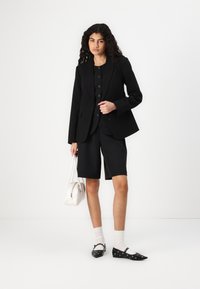 Blazer noir ajusté sur une chemise à boutons assortie et un short noir. Les accessoires incluent un sac à main blanc et des ballerines noires à motifs.