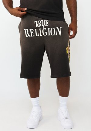 Uomo con pantaloncini neri True Religion, calzini bianchi e sneakers bianchi, che solleva leggermente la maglietta nera per mostrare la vita.