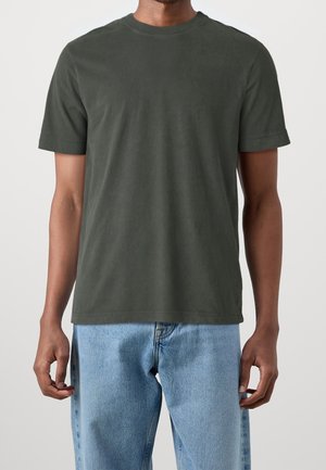 Homme portant un t-shirt manches courtes vert foncé et un jean bleu clair, debout les bras détendus le long du corps sur un fond uni.
