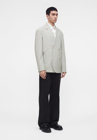 Homme debout portant un blazer croisé gris clair, une chemise blanche, un pantalon noir à jambes larges et des chaussures noires sur un fond uni.