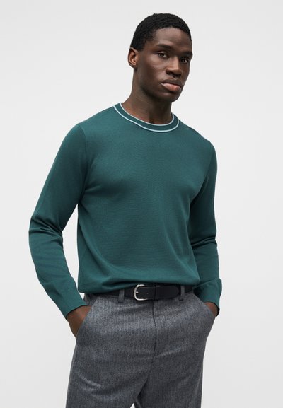 Πράσινο πλεκτό πουλόβερ με στρογγυλή λαιμόκοψη και αντίθεση σε ανοιχτό μπλε, συνδυασμένο με γκρι παντελόνι με μοτίβο και μαύρη ζώνη.