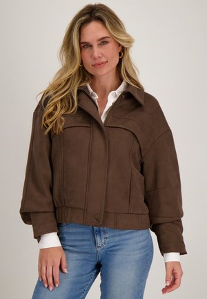 Vrouw met lang blond haar, draagt gouden oorringen, een bruin jack met ritssluiting over een wit shirt en blauwe spijkerbroek, staand tegen een effen achtergrond.