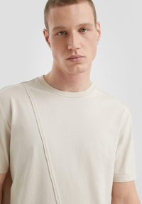 Beżowa bawełniana koszulka z okrągłym dekoltem i krótkim rękawem, z teksturowanym diagonalnym przeszyciem na przodzie. Gładka powierzchnia materiału.