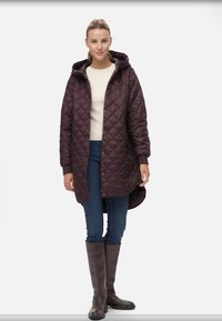 Quiltad mörkbrun kappa med huva, tryckknappar och sidofickor, bärs över en krämfärgad tröja och blå jeans, med höga bruna stövlar.