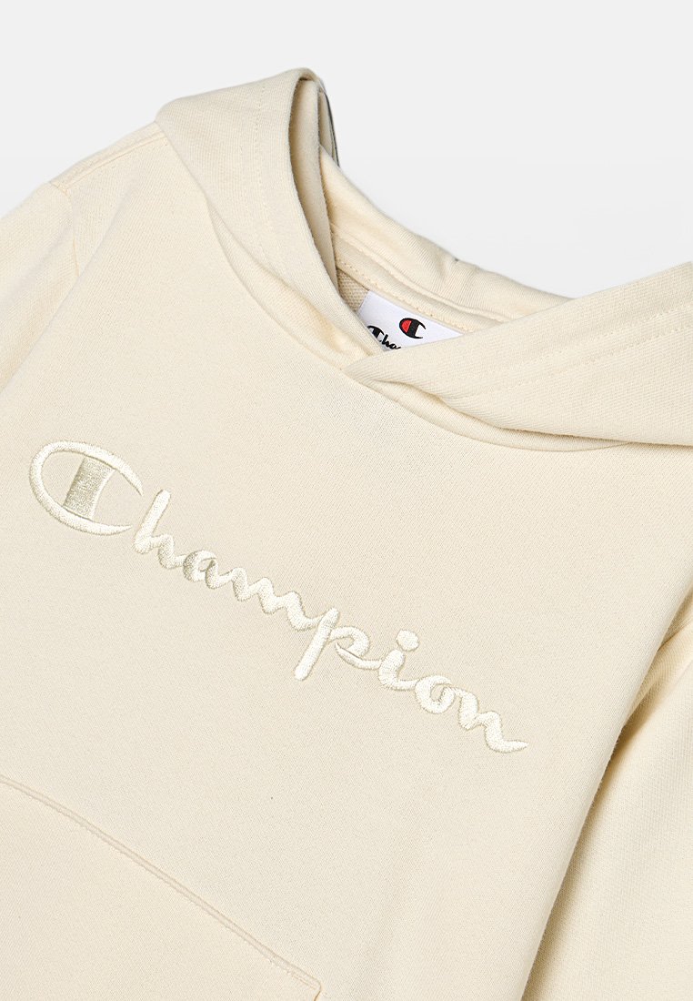 Sudadera color crema hecha de un tejido suave que presenta un logo bordado de "Champion" en blanco, con capucha ajustable y bolsillo frontal.