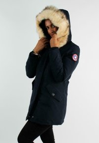 Parka d'hiver en marine avec une capuche bordée de fourrure, des poches avant et des fermetures à boutons. Présente un patch logo sur la manche. Texture douce.