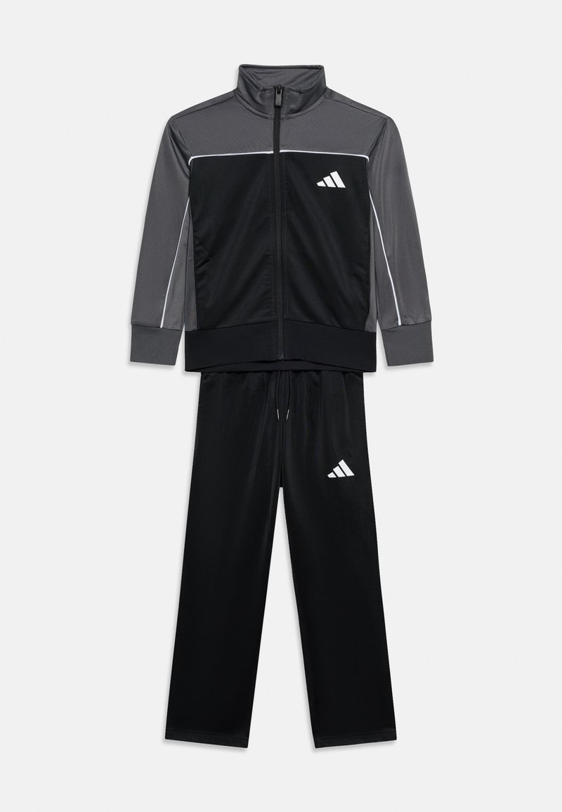 Αθλητική φόρμα δύο τεμαχίων με μαύρο παντελόνι και μαύρο-γκρι μπουφάν με φερμουάρ, με λευκό λογότυπο στο στήθος και τον μηρό.