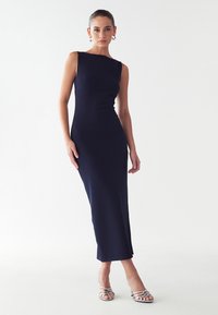 Jūras zila bezpiedurkņu kleita, šaura piegriezuma, līdz ceļiem, ar taisnu apakšmalu. Valkāta ar sudraba siksnu papēžiem. Gluda auduma tekstūra.