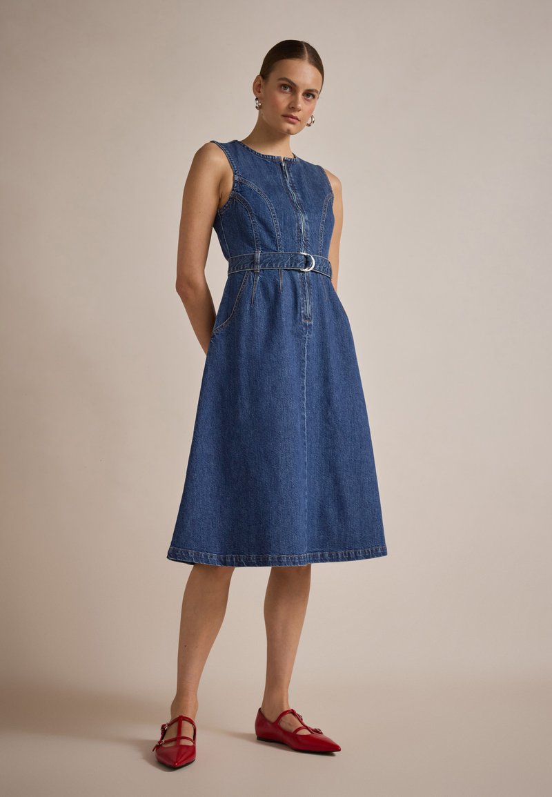 Denim A-linjainen mekko, jossa on olkapäätön suunnittelu, etuvetoketju, muotoiltu vyötärö ja yhteensopiva vyö. Omistaa hienovaraisen rakenteen ja sinisen värin. Punaiset kengät korostavat.