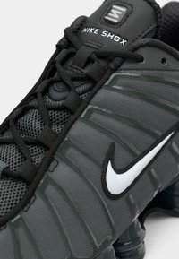Nike Shox sportbačiai pilkos spalvos su tekstūruotu tinkliniu paviršiumi, plačiomis raištelėmis, tamsiais akcentais, išryškintu swoosh logotipu ir paminkštintu guminio pado sole.