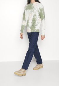 Sudadera tie-dye en verde claro y beige, combinada con jeans de mezclilla azules y botas de senderismo beige con herrajes plateados. Textura suave.