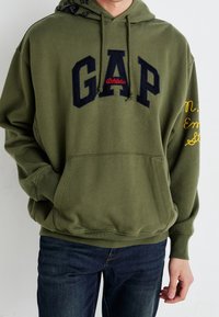 Sweatshirt à capuche vert avec lelettrage "GAP" en marine et un détail "Athletic" rouge. Comprend une poche avant et un texte brodé jaune sur la manche.