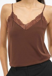 Brązowy top na cienkich, regulowanych ramiączkach z koronkową dekoracją przy dekolcie w kształcie litery V, wykonany z miękkiego, lejącego się materiału. W zestawie z czarnymi spodniami.