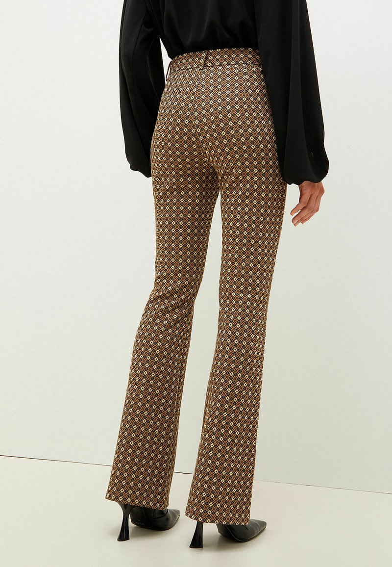 Osoba ubrana w brązowe i czarne wzorzyste spodnie z wysokim stanem, czarną bluzkę oraz czarne buty na obcasie, stojąca na tle jednolitym.