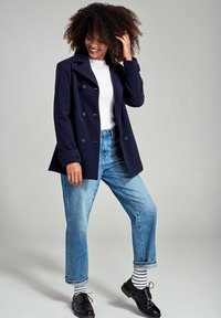 Cappotto navy doppiopetto con grandi bottoni, indossato sopra un maglione bianco e jeans blu, abbinato a scarpe nere e calzini a righe.