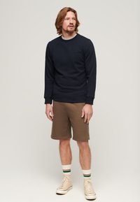 Felpa navy in cotone con scollo rotondo e polsini a coste, abbinata a pantaloni corti marroni. Sneakers beige con lacci bianchi e calze a righe.