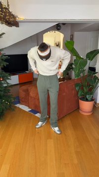 Gray ribbed sweater with logo, olive green sweatpants, and silver shoes. Indoor setting with a couch and potted plant in the background.