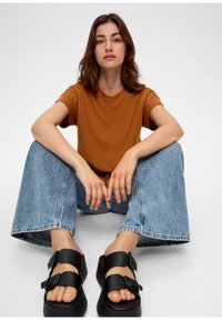 Hnědé tričko s krátkým rukávem, světle modré široké džíny s třepením na lemu a černé sandály se dvěma přezkami, sedící na neutrálním pozadí.