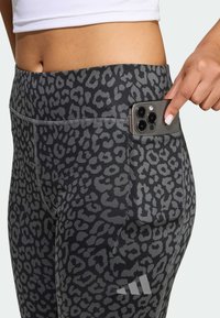 Čierne a sivé legíny so vzorom leopardích škvŕn, vybavené bočným vreckom na smartfón, prešívanými detailmi a logom.