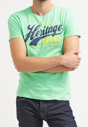 T-shirt en coton vert avec un col rond, affichant un large texte graphique "Heritage Brand" en bleu marine et jaune sur le devant. Manches courtes.