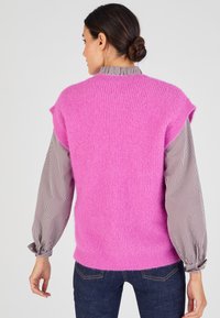 Fluffy roze mouwloze pullover met een ribbelstructuur, gelaagd over een gestreept shirt met lange mouwen. Donkere jeans met subtiele stiksels zichtbaar.