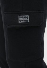 Sorte sweatpants med en stor sidelomme og et stofmærke med teksten "Pier One" med hvide syninger. Blødt, tekstureret materiale.