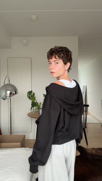 Schwarze Zip-Up-Hoodie mit lockerer Passform, getragen über einem weißen ärmellosen Top. Hellgraue Jogginghose, minimaler Textur. Innenraumszene mit Pflanzen.