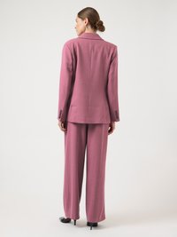 Femme debout de dos portant un blazer mauve ajusté et un pantalon large assorti avec des talons noirs, sur un fond blanc uni.