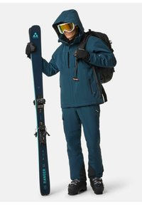 Tannengrüne Skijacke mit Kapuze, dazu passende Hose und Handschuhe. Hält einen blauen Ski mit schwarzen Bindungen. Ausgestattet mit Skibrille und einem schwarzen Rucksack.