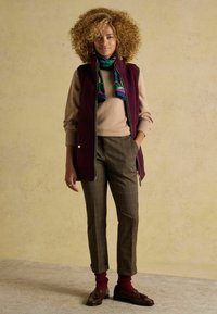 Sarkans rāvējslēdzēja bezpiedurkņu veste, virs pelēka džempera, kombinēta ar brūnām raibām biksēm un brūniem loaferi. Aksesuārs - krāsains zīda šalle.