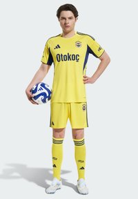 Žltý futbalový dres s námorníckymi akcentmi, obsahujúci logo 'Otokoc' a tri pruhy na rukávoch. Hráč drží modro-biely futbal.