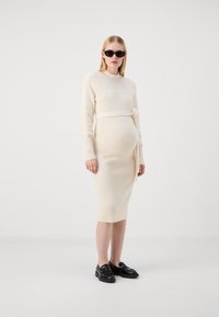 Abito premaman in maglia crema con maniche lunghe, silhouette aderente e dettaglio in vita legato, abbinato a mocassini neri e occhiali da sole.