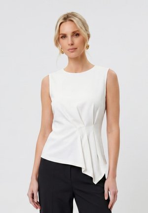 Femme portant un top asymétrique blanc sans manches avec des plis, associé à un pantalon noir et des boucles d'oreilles dorées sur un fond clair uni.
