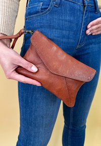Clutch in pelle marrone con finitura testurizzata, dotato di una patta triangolare e di una tracolla staccabile, tenuto contro dei jeans blu.