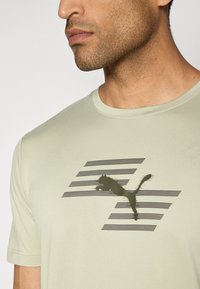 Lys grønn treningsskjorte laget av mykt materiale, med en svart Puma-logo og horisontale striper i bakgrunnen. Korte ermer, rund krage.