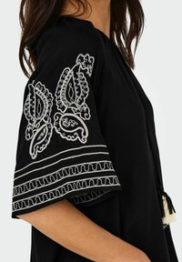 Vêtement noir avec manches courtes brodées. Présente des motifs cachemire blancs et des bordures décoratives. Accents en pompons à l'ourlet.