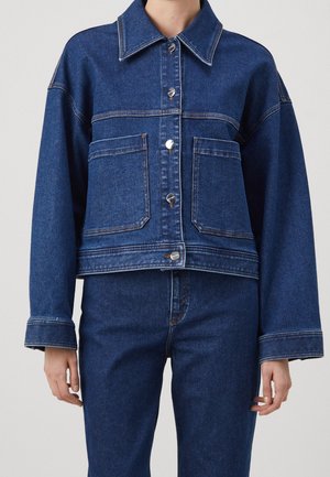 Un mannequin porte une veste en jean bleu foncé à col large, avec des boutons argentés et deux grandes poches avant, assortie à un jean.