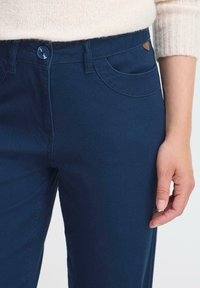 Detailní záběr na osobu v námořnický modrých kalhotách a krémovém pleteném svetru, který zobrazuje knoflík, kapsy a pravou ruku po boku.