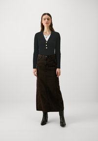Haut long à manches longues à rayures noires avec des boutons dorés, associé à une jupe maxi en denim marron et des bottes à talons noires. Collier en couches simple visible.