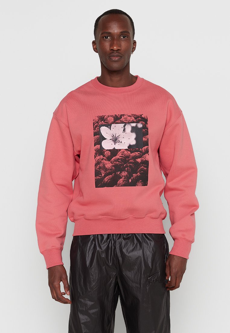 Mężczyzna w koralowej bluzie z czarno-białą grafiką kwiatu i błyszczących czarnych spodniach Nike, stojący na jednolitym jasnym tle.